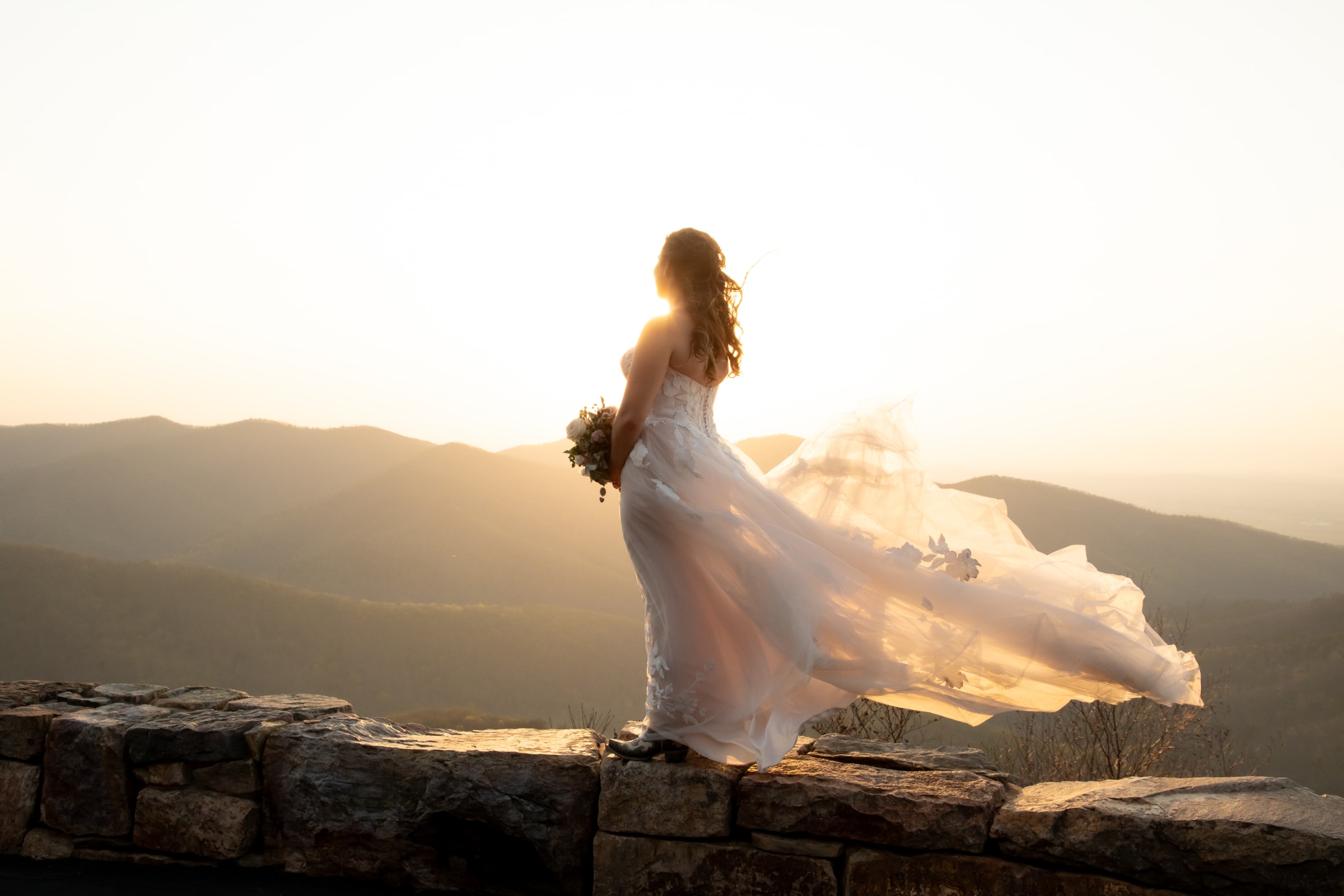 Bride in flowing white gown on a stone overlook at golden hour, Blue Ridge Mountains behind her, dress billowing in the wind