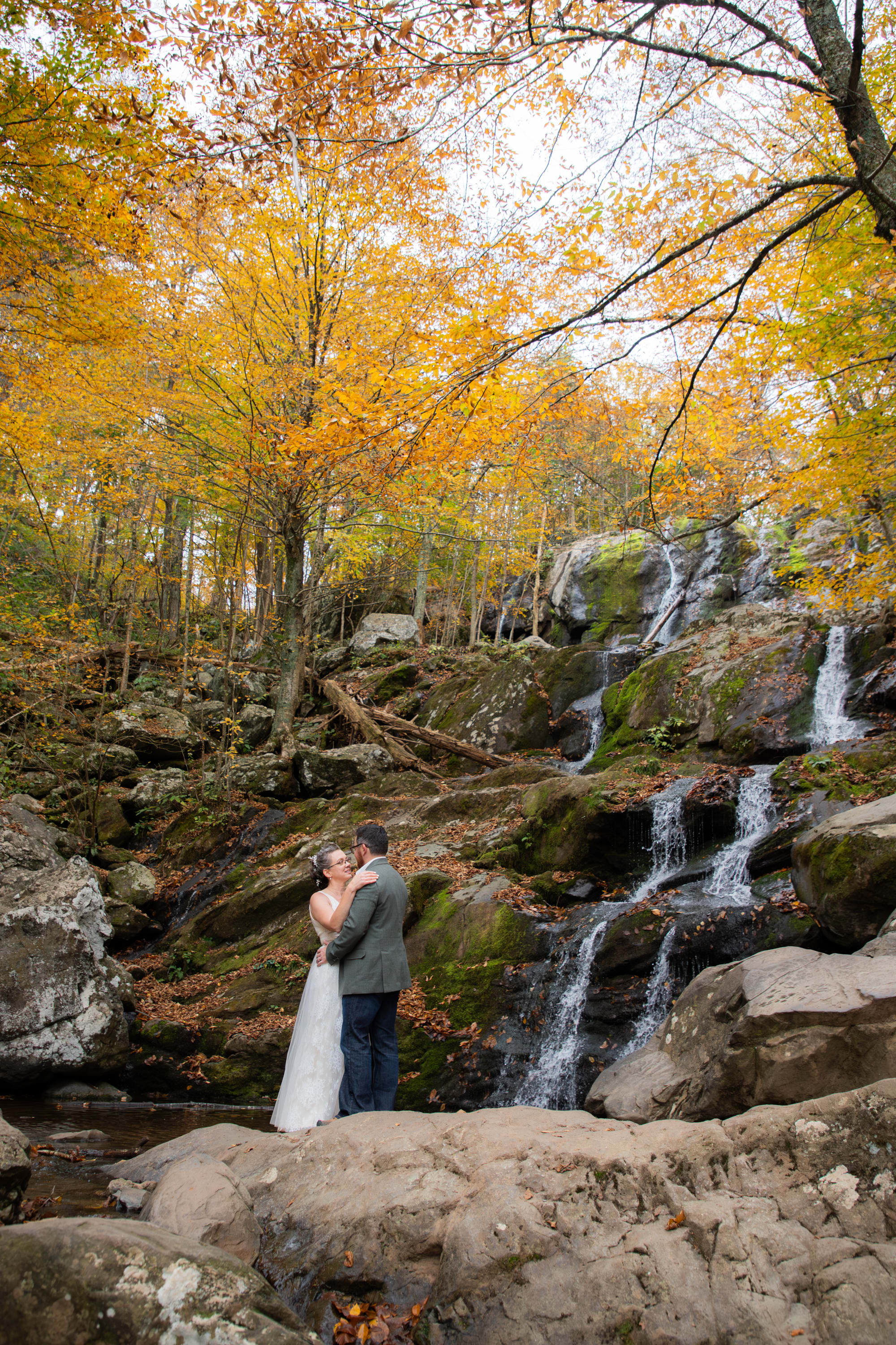 Couple embracing at Dark Hollow Falls in fall with golden foliage and tiered waterfall behind them