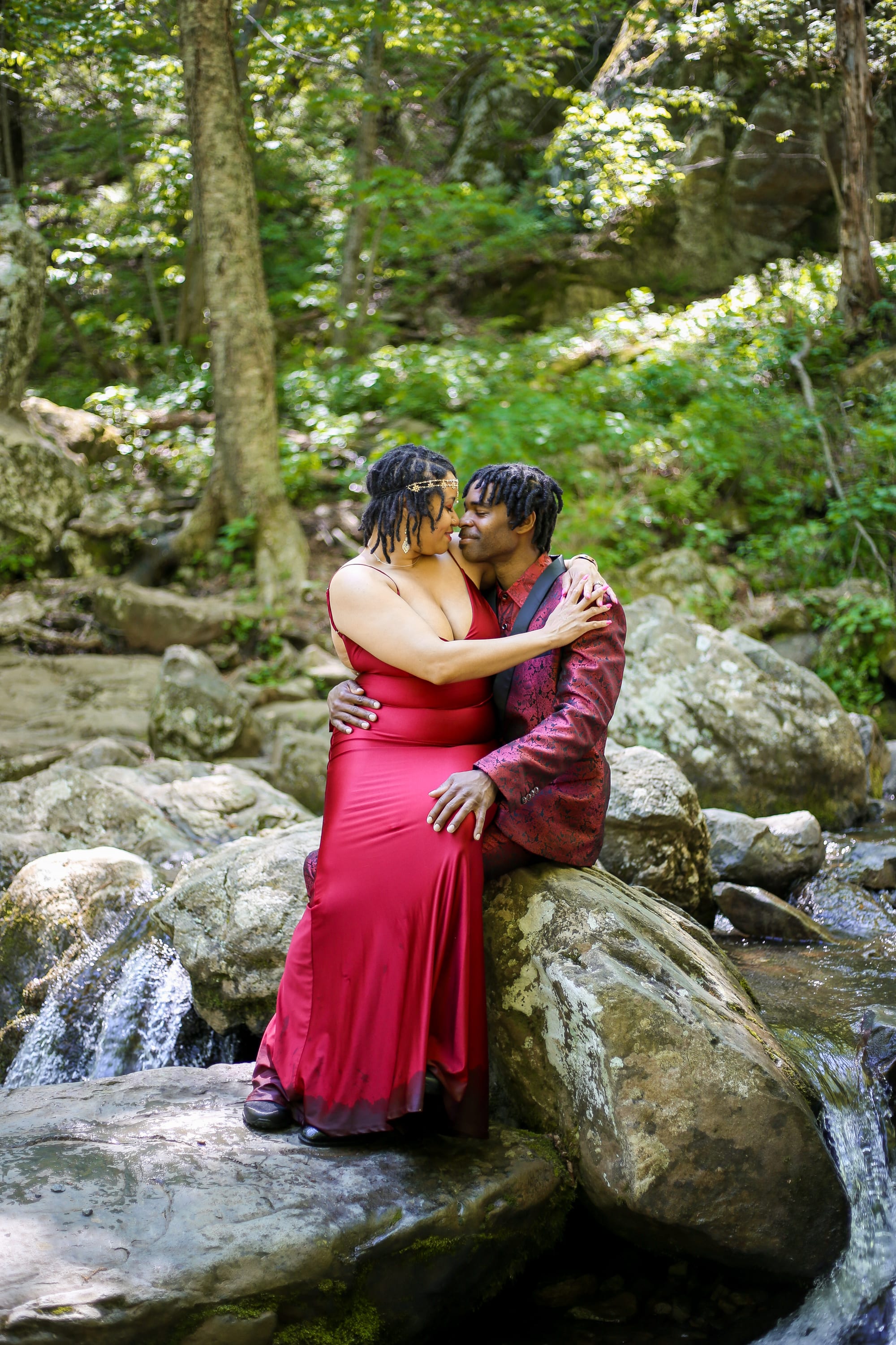 Couple in a red dress embracing on the rocks at the base of a waterfall, surrounded by lush green summer forest in Shenandoah National Park