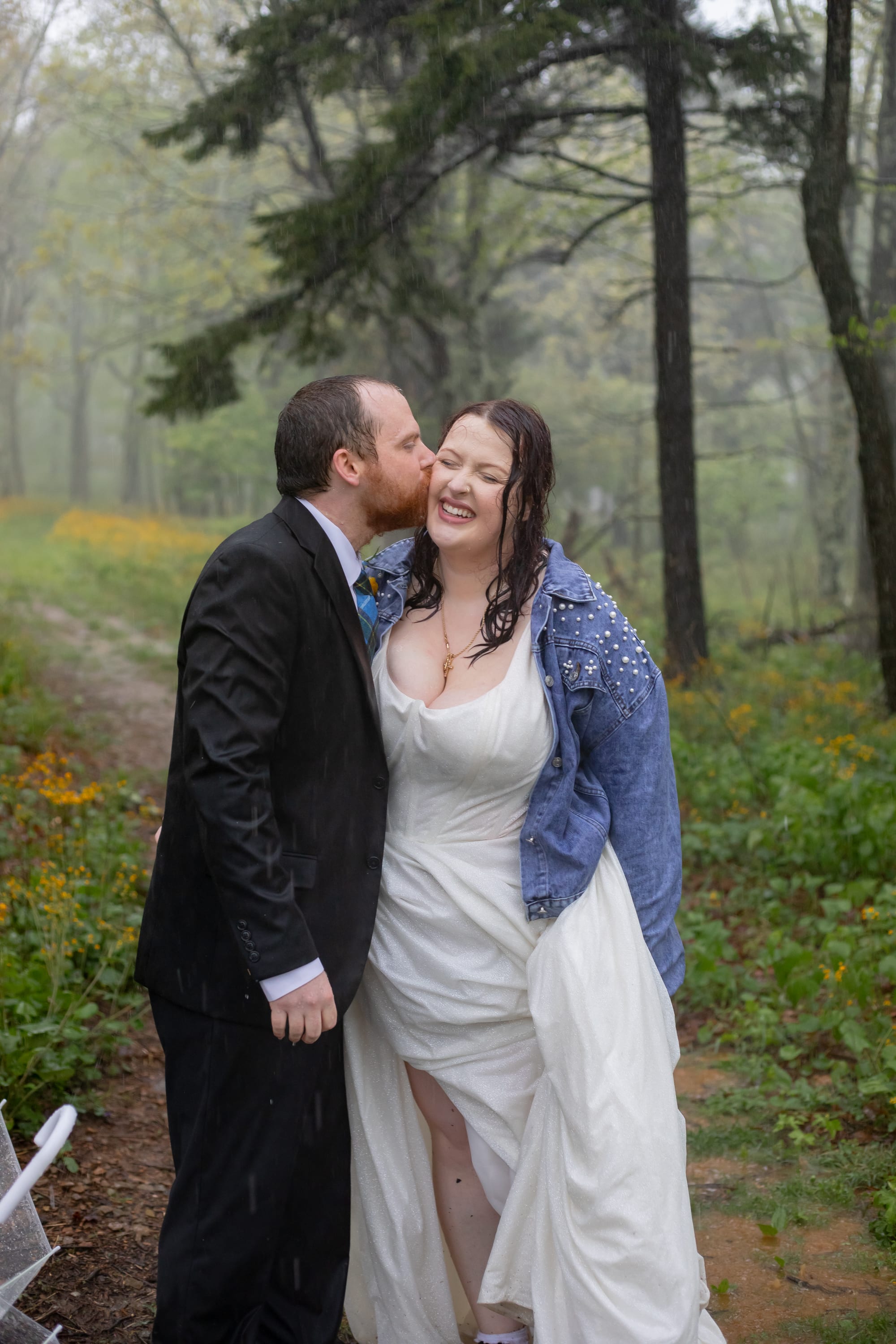 Groom kissing bride on the cheek in a misty Shenandoah forest, bride laughing in a pearl denim jacket with yellow wildflowers behind them