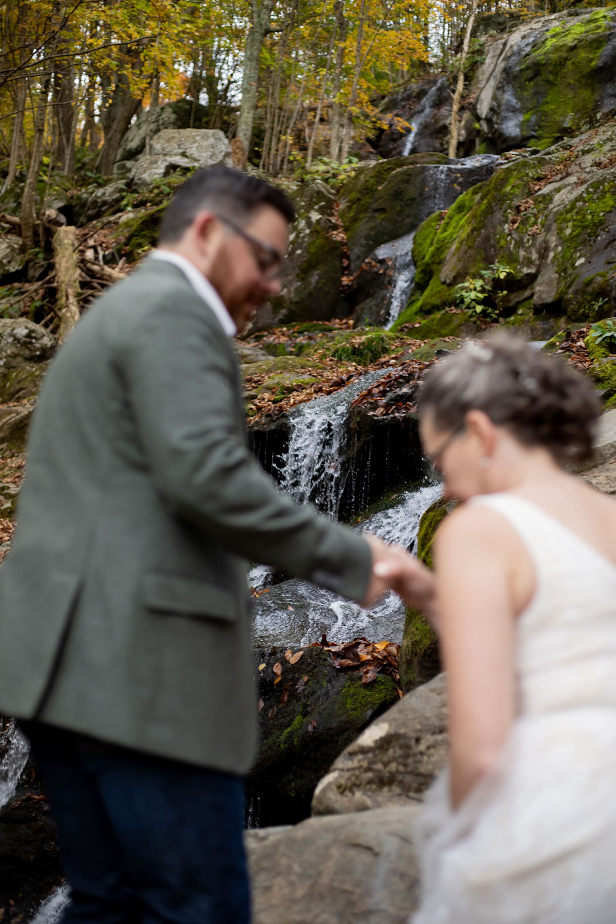 Groom helping bride over rocks at a waterfall in Shenandoah National Park during fall foliage
