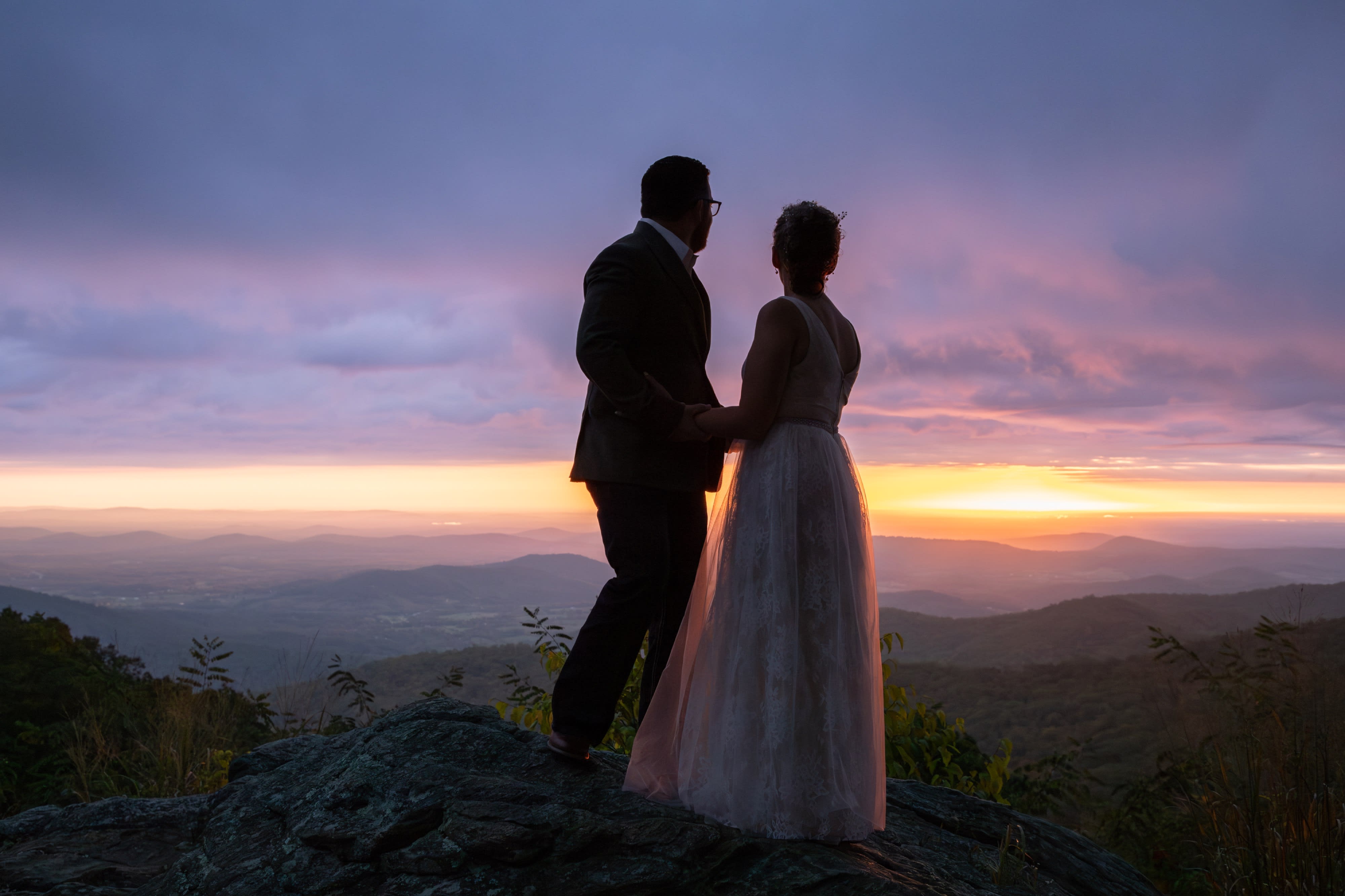 Couple standing on a rocky Shenandoah summit, backs to camera, watching a vivid pink and magenta sunset sky stretch across the Blue Ridge Mountains