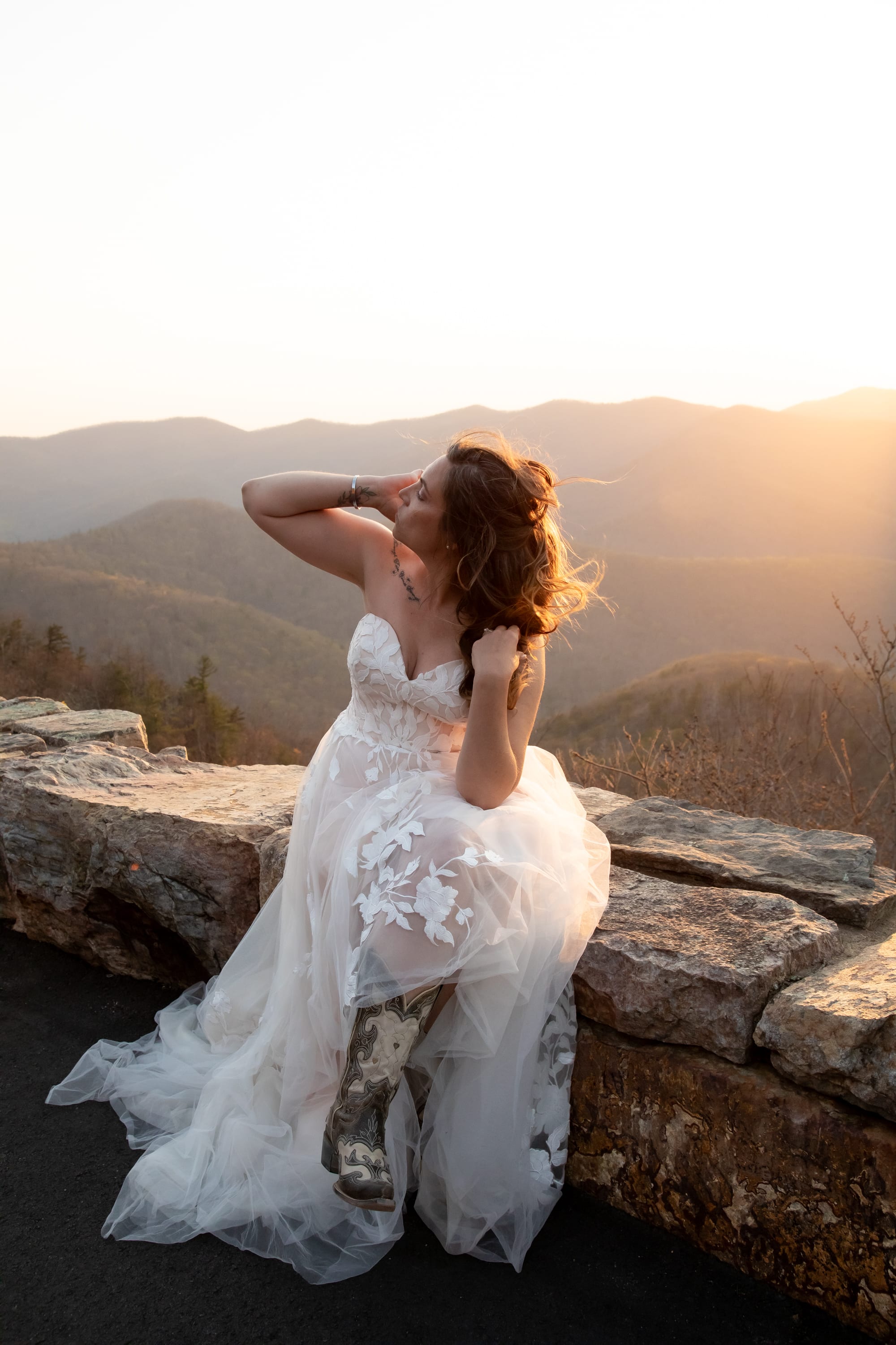 Bride in white lace gown and cowboy boots sitting on a stone overlook wall, head tilted back in the wind, golden Blue Ridge ridges behind her at sunset