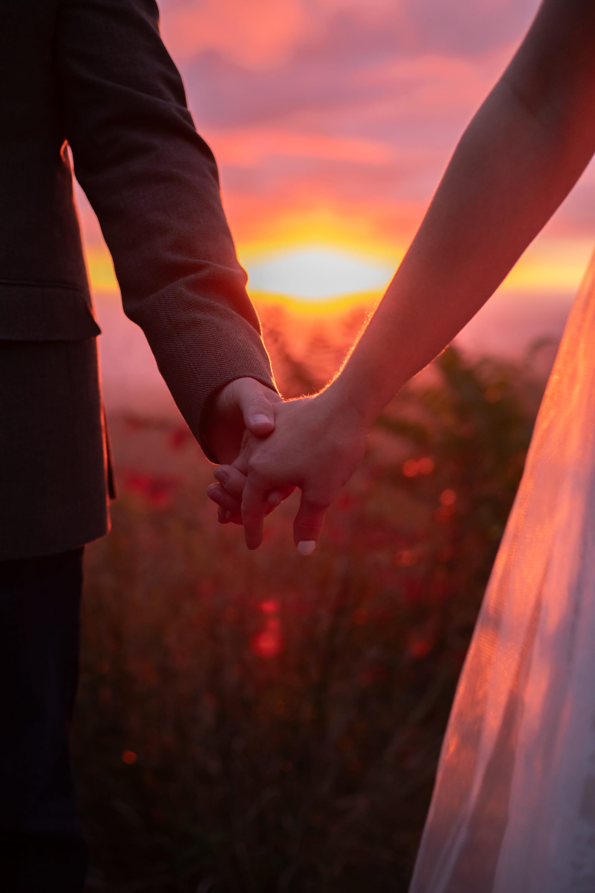 Close-up of couple holding hands with a fiery red and orange sunset sky glowing behind them, veil catching the light