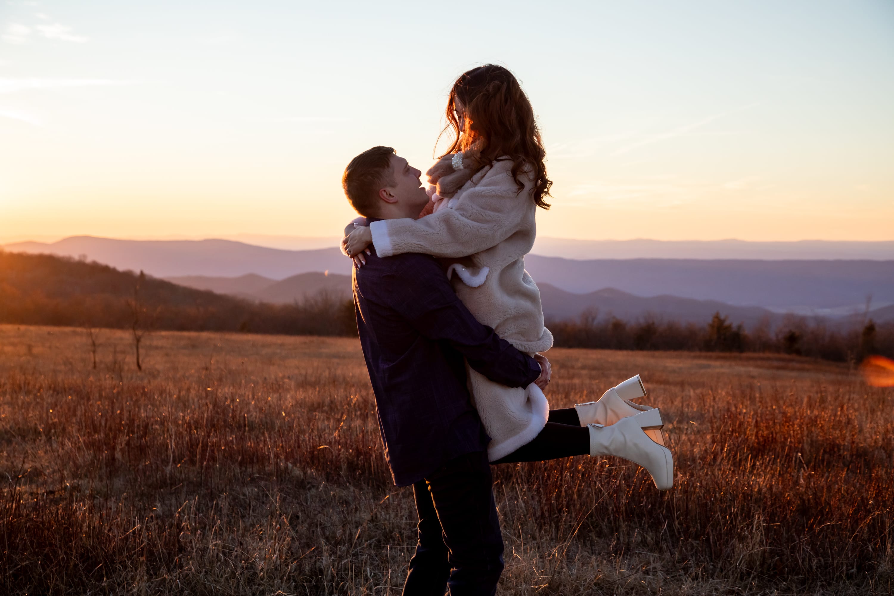 Groom lifting bride in a golden open meadow at sunset with layered Blue Ridge Mountain ridges stretching across the horizon behind them