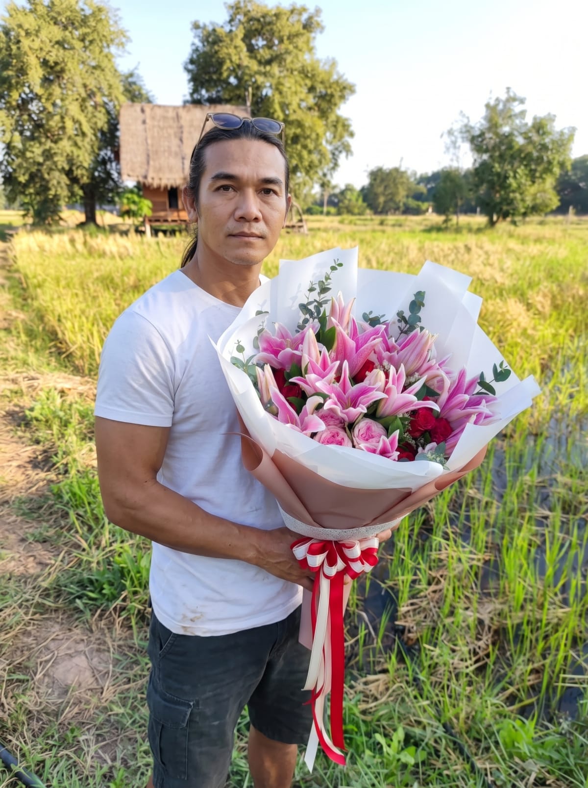 The shop owner holding a pink lily bouquet in a rice field, with a thatched hut behind him
