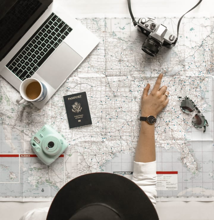 Hand pointing to a map beside a passport, camera, laptop and coffee while planning a trip.