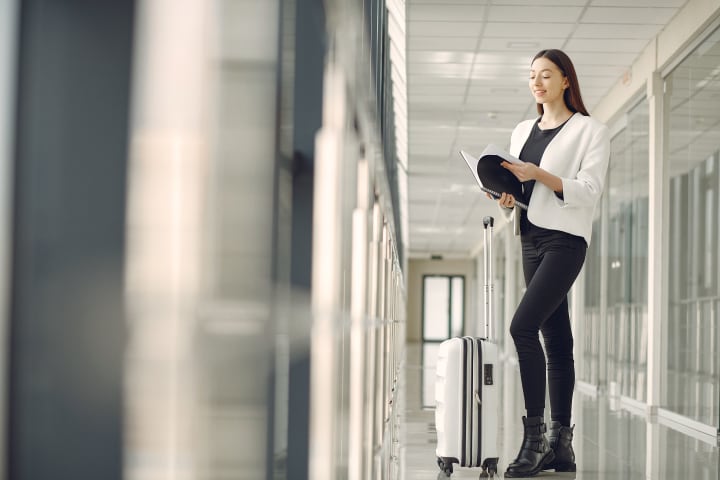 Full body of slim female businesswoman in formal clothes with suitcase standing in airport hallway and reading note in notebook while waiting for flight