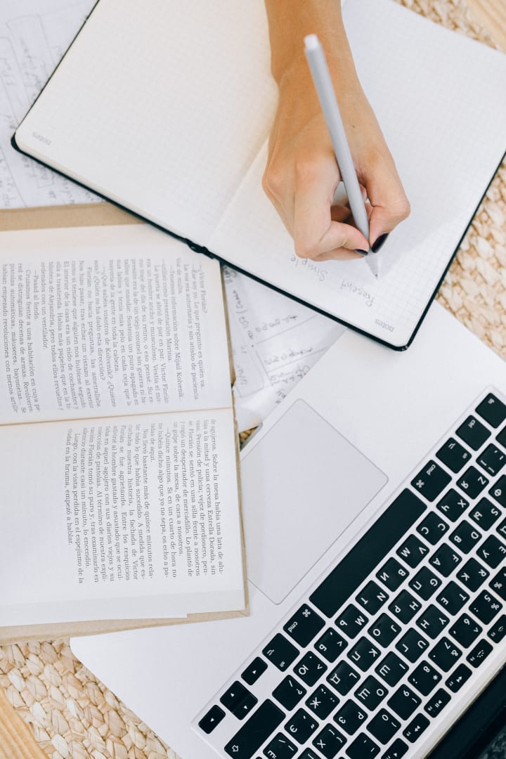 Person writing in a notebook beside an open laptop and books.