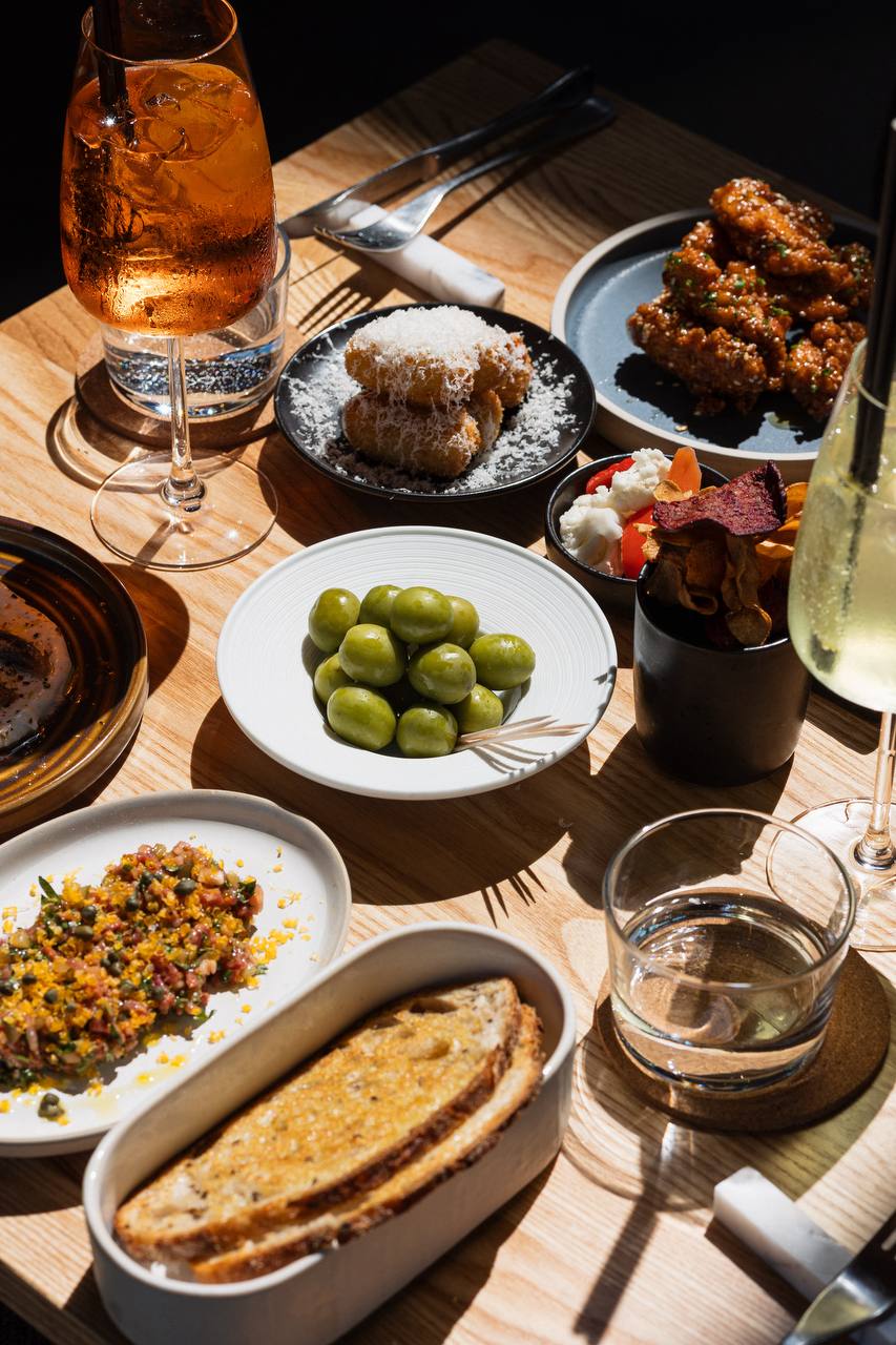 An overhead shot of assorted small plates and snacks at Cafe Calton, featuring green olives, fried items, and a drink, bathed in natural light
