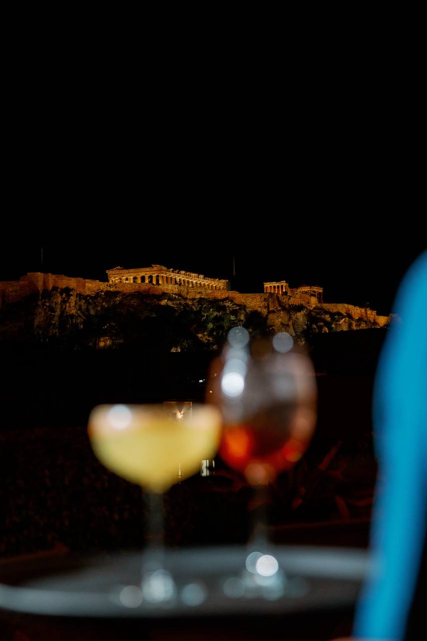 Two cocktail glasses on a table with the illuminated Parthenon and Acropolis visible in the background at night from Retiré Bar