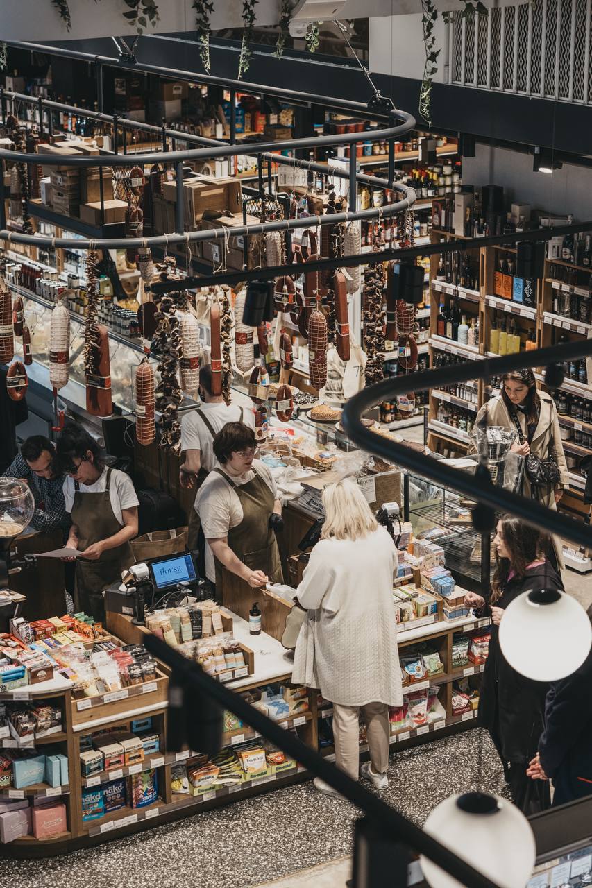 Overhead view of the ERGON House Athens Agora where apron-clad staff assist customers among shelves of gourmet products and hanging cured meats