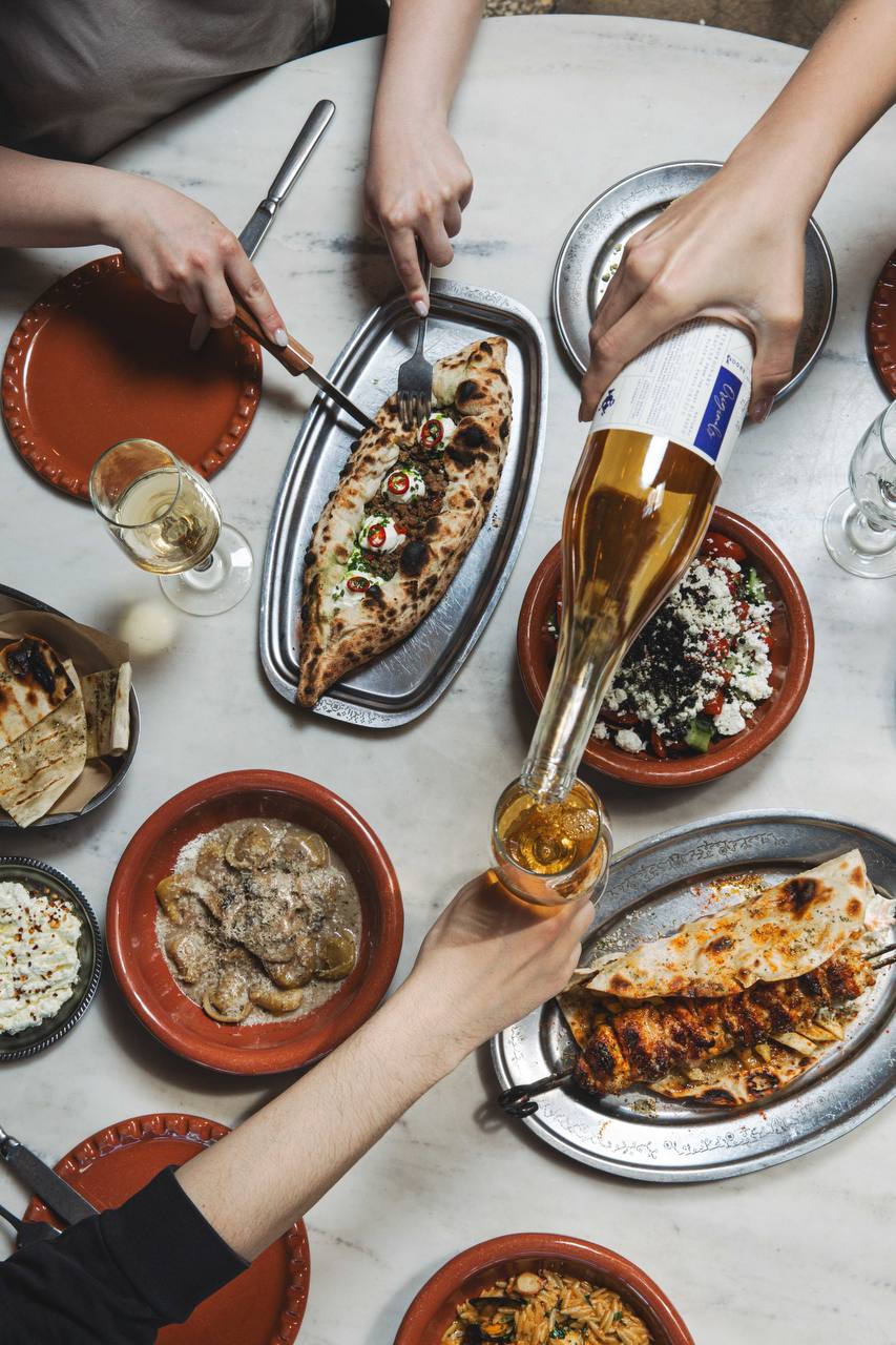 Hands sharing dishes and pouring wine at a marble table in the communal dining area at ERGON House Athens