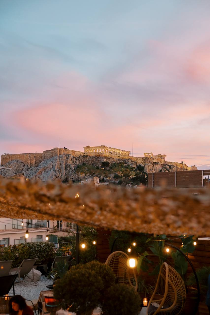 Rooftop terrace at Retiré Bar with string lights and wicker chairs framing a sunset view of the Acropolis and Parthenon