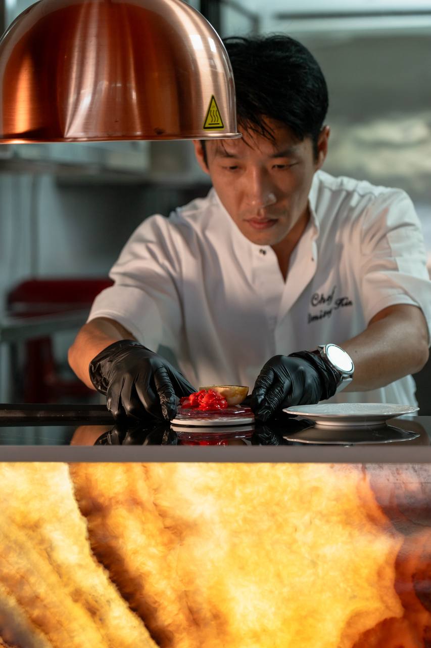 Chef Dmitry Kim plating a dish under a copper heat lamp in LUNA's open kitchen, Koh Samui