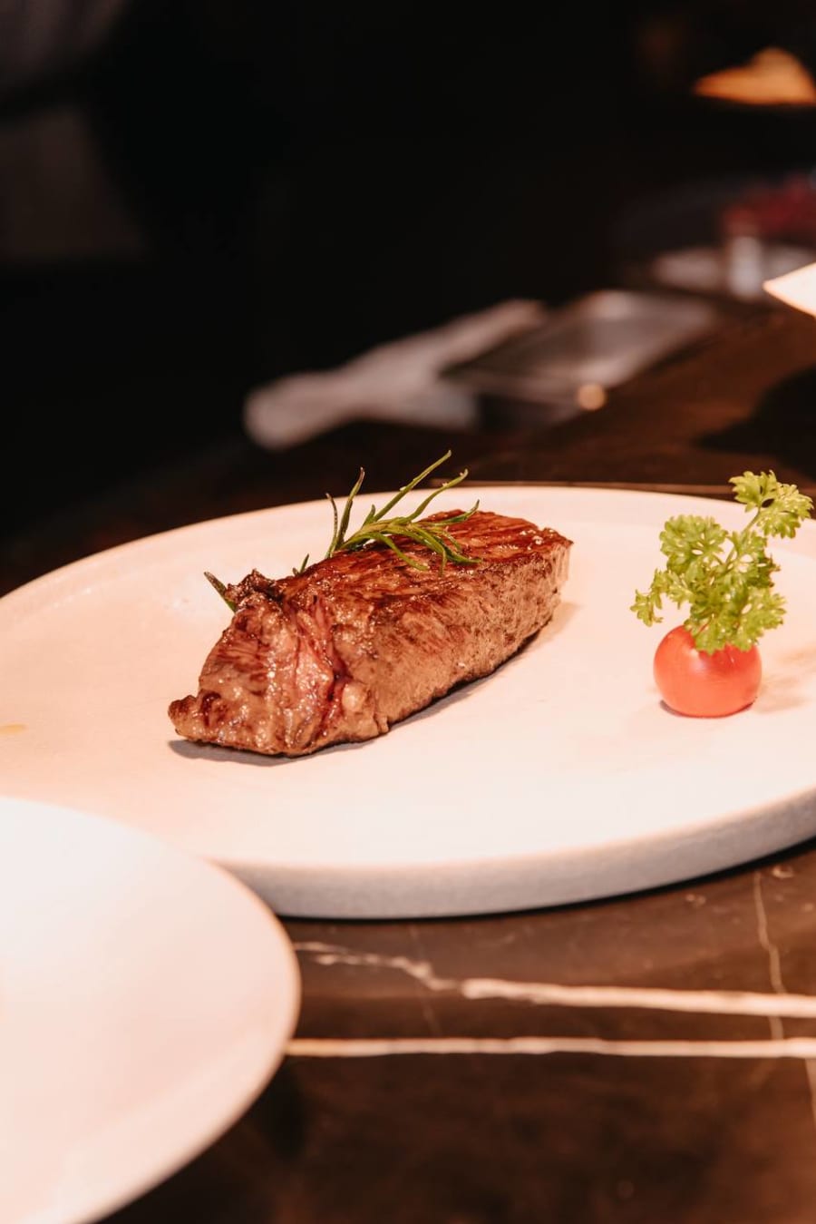 Perfectly seared dry-aged steak on white plate with rosemary sprig on dark marble at PRIME Saigon