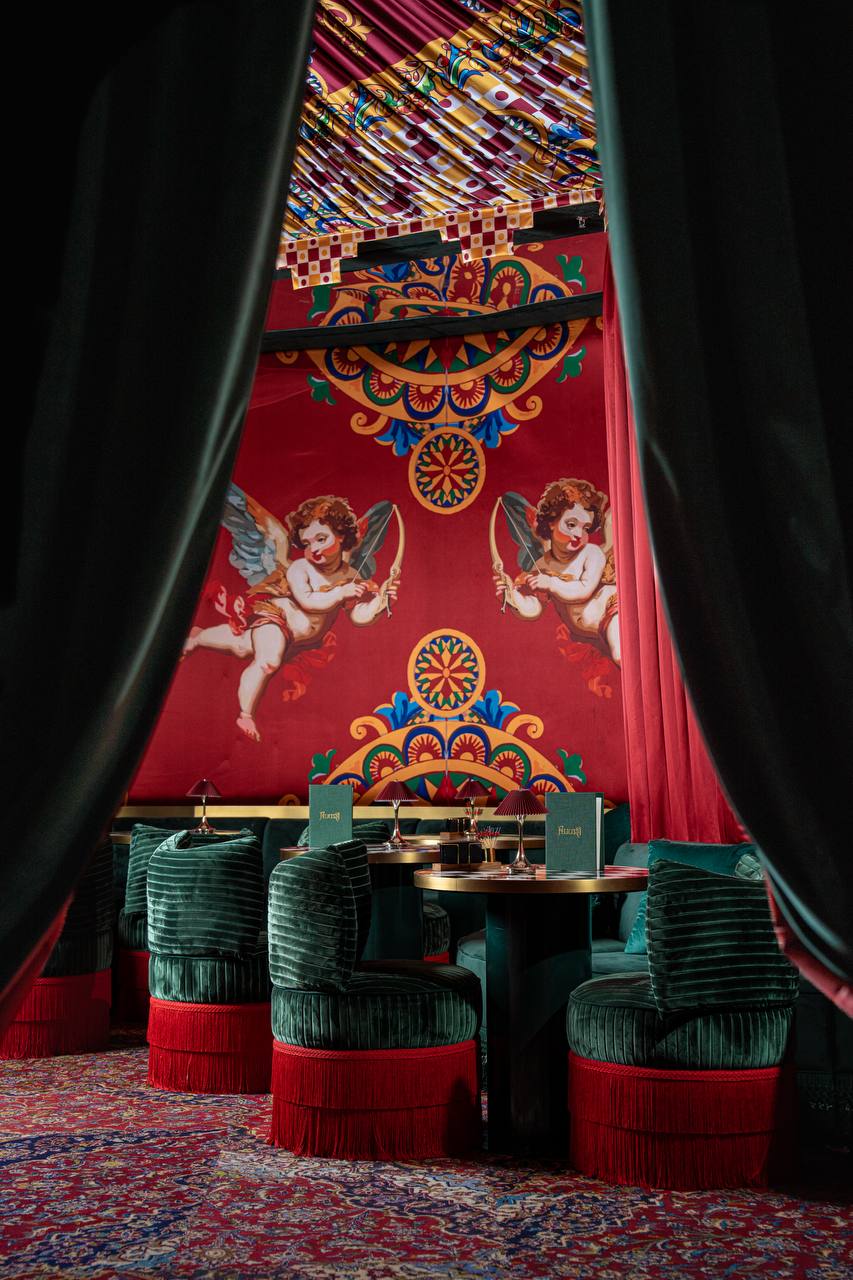 Velvet curtains reveal a maximalist dining room with cherub murals and emerald seating at Felicità