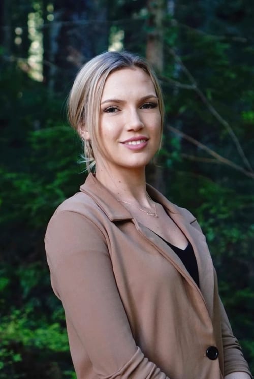 Professionally dressed woman in a beige blazer smiling outdoors in a dense forest.