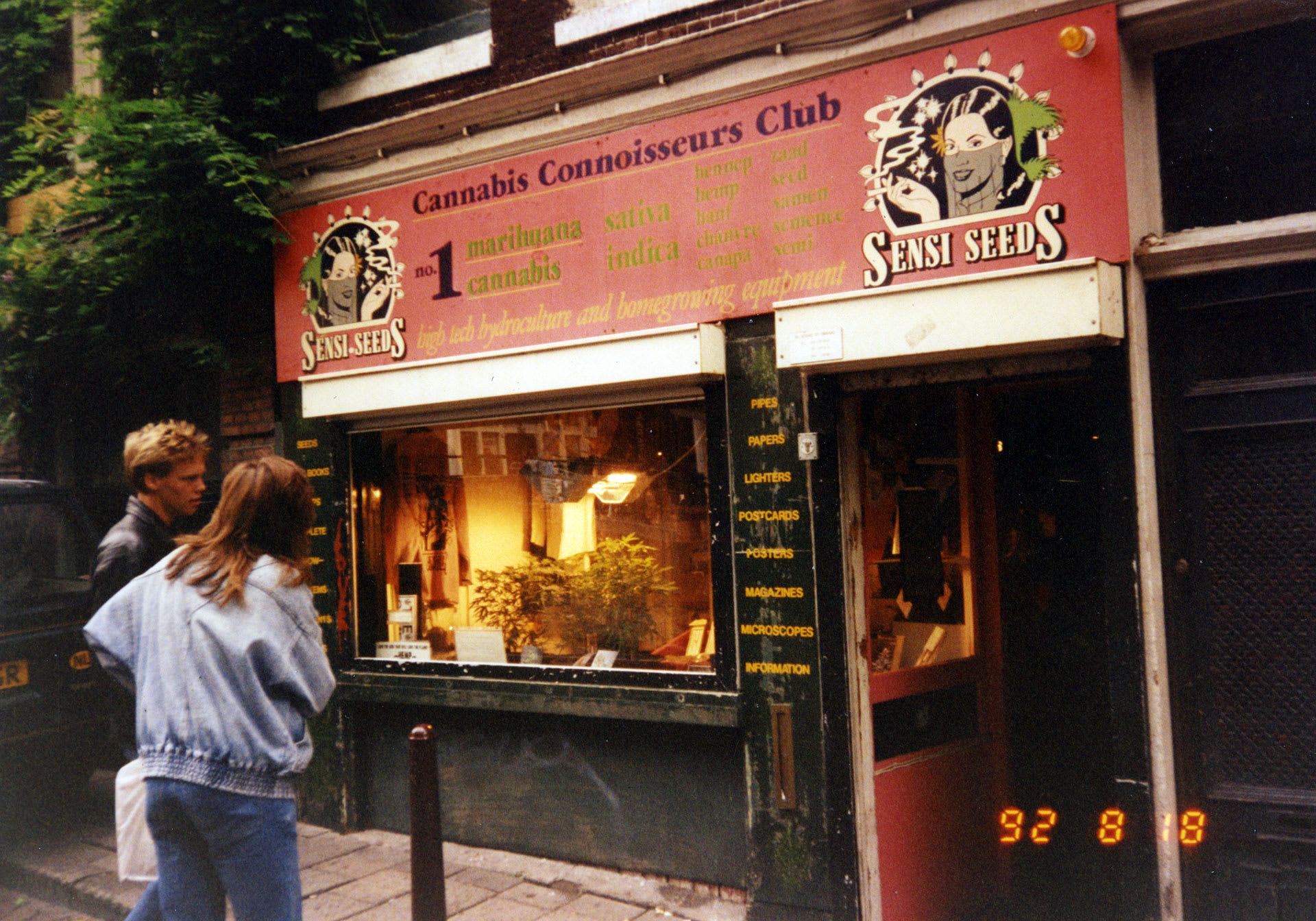 Storefront of the Cannabis Connoisseurs Club with a Sensi Seeds sign and passersby outside.