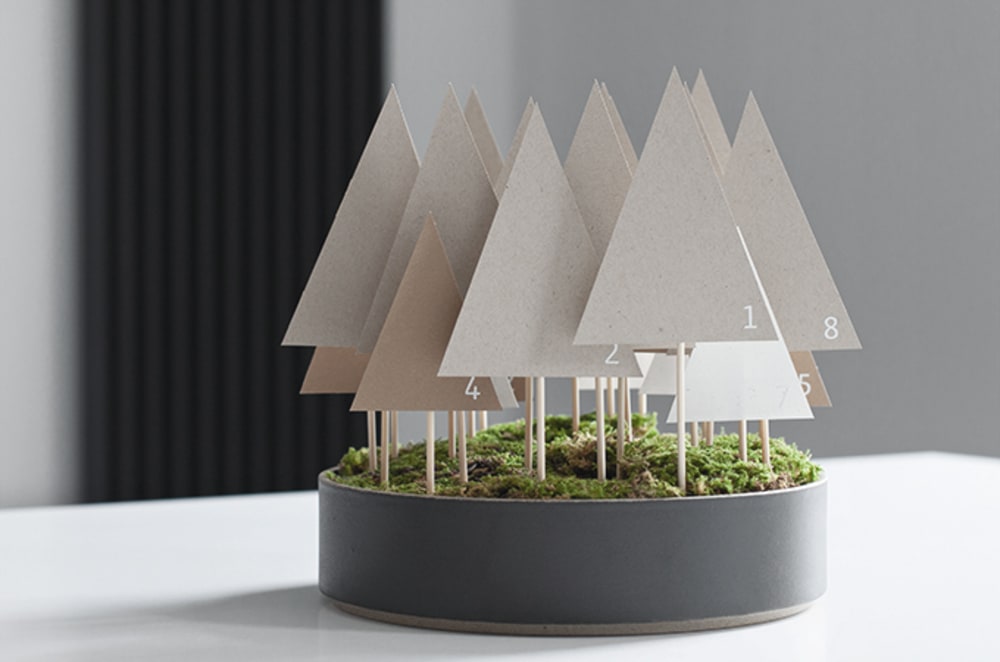 Advent calendar made of paper trees mounted on a wooden stick and tucked in a container onto a moss base.<span class="sr-only"> (opened in a new window/tab)</span>