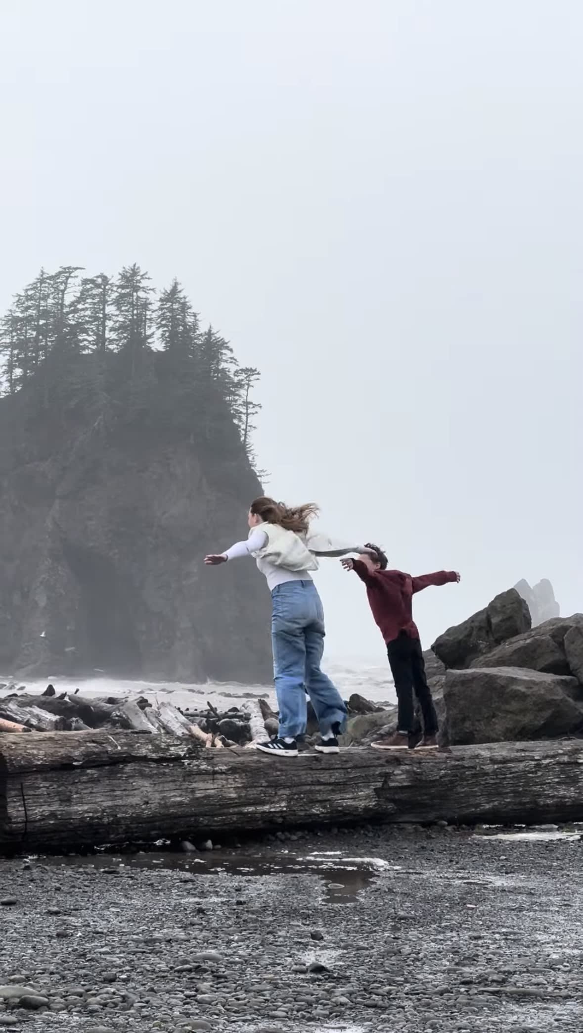 Kids with arms outstretched on a driftwood log with misty sea stacks behind at La Push, Washington