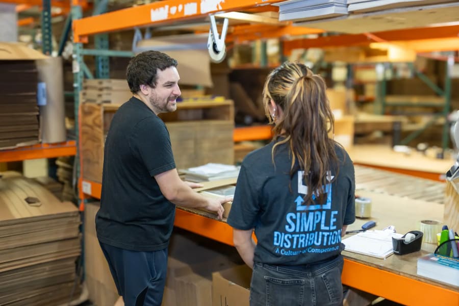 Two Simple Distribution team members talking face to face at a workbench on the warehouse floor
