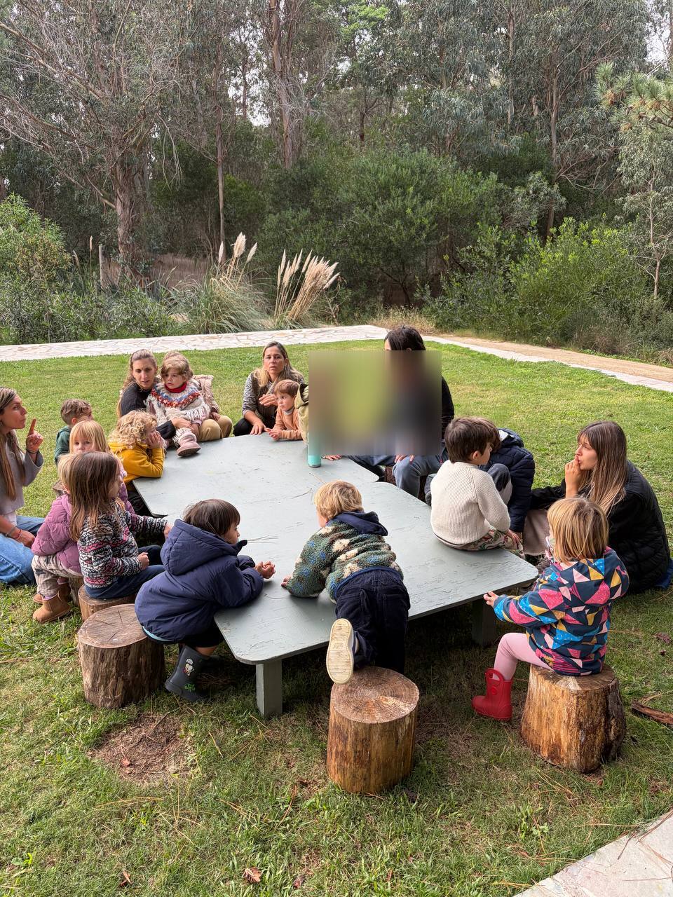 Niños, niñas y adultas reunidos alrededor de la mesa exterior