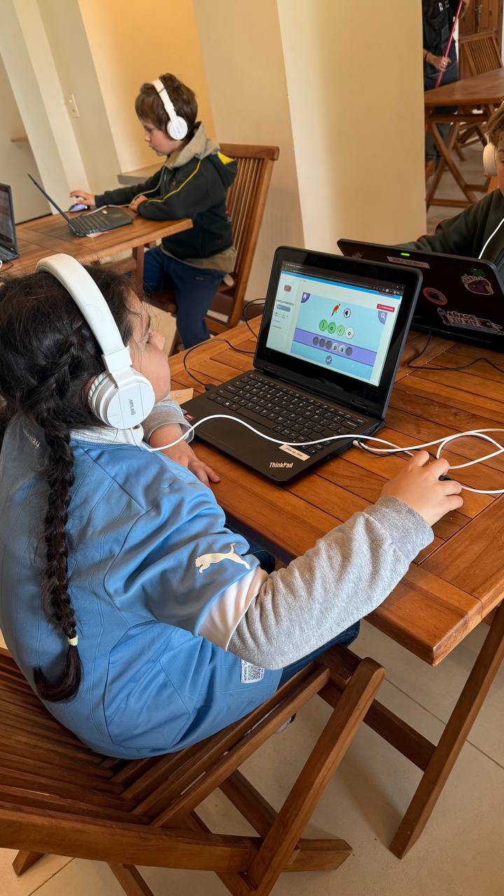 Niños y niñas realizan juegos de lectoescritura en una plataforma digital personalizada, con auriculares y acompañamiento adulto cercano durante la actividad.