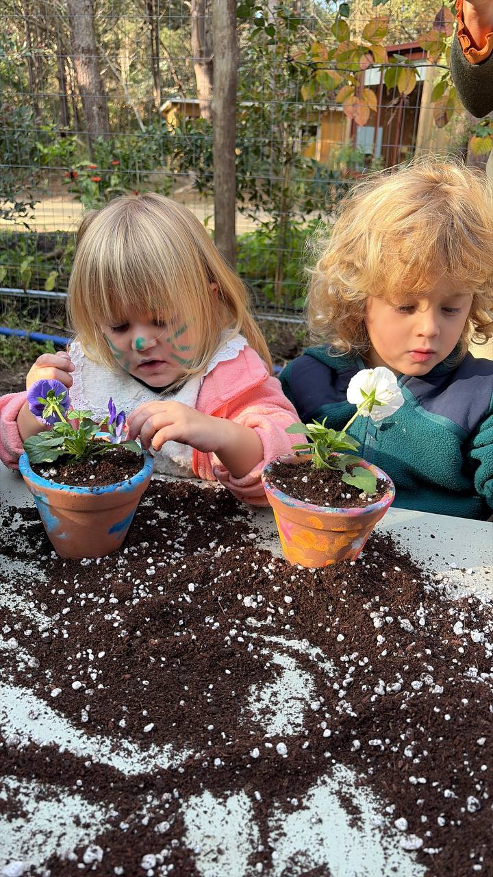 Dos ninos plantando pensamientos en macetas decoradas sobre una mesa con tierra