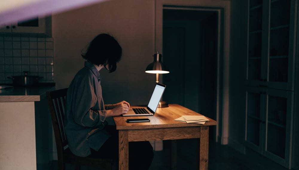 A person sitting alone at a kitchen table late at night, laptop open and glowing, phone face down beside a small stack o