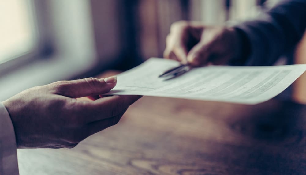 Two hands across a wooden desk, one passing a printed document to the other, a pen resting on the page, soft daylight fr