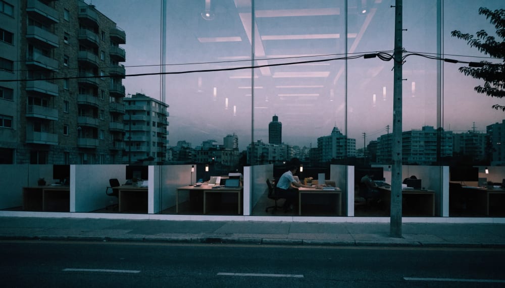 An open plan Tel Aviv office seen from the street at dusk through a glass facade, a handful of desks still occupied, the