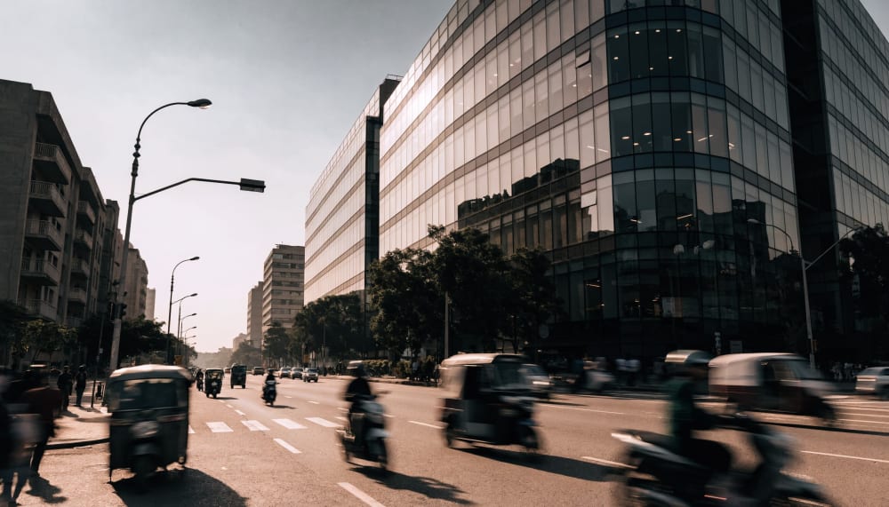A wide daylight view of a modern glass office tower in a busy South Asian tech district, motorbikes and auto rickshaws b