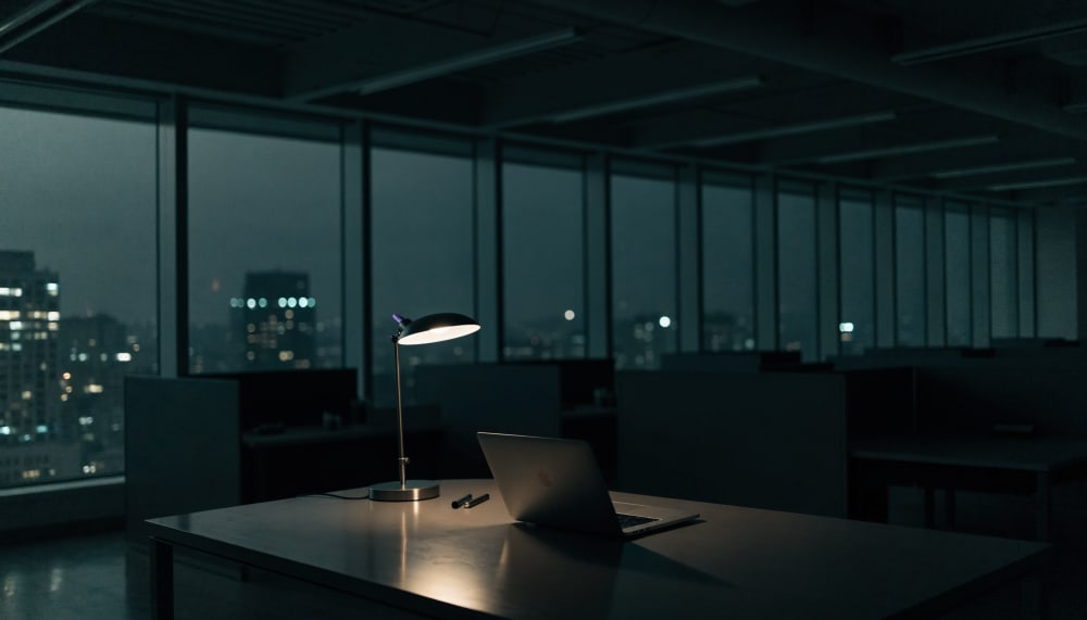 A single desk lamp lit at night in an empty open plan office, a laptop open showing an abstract terminal interface, city