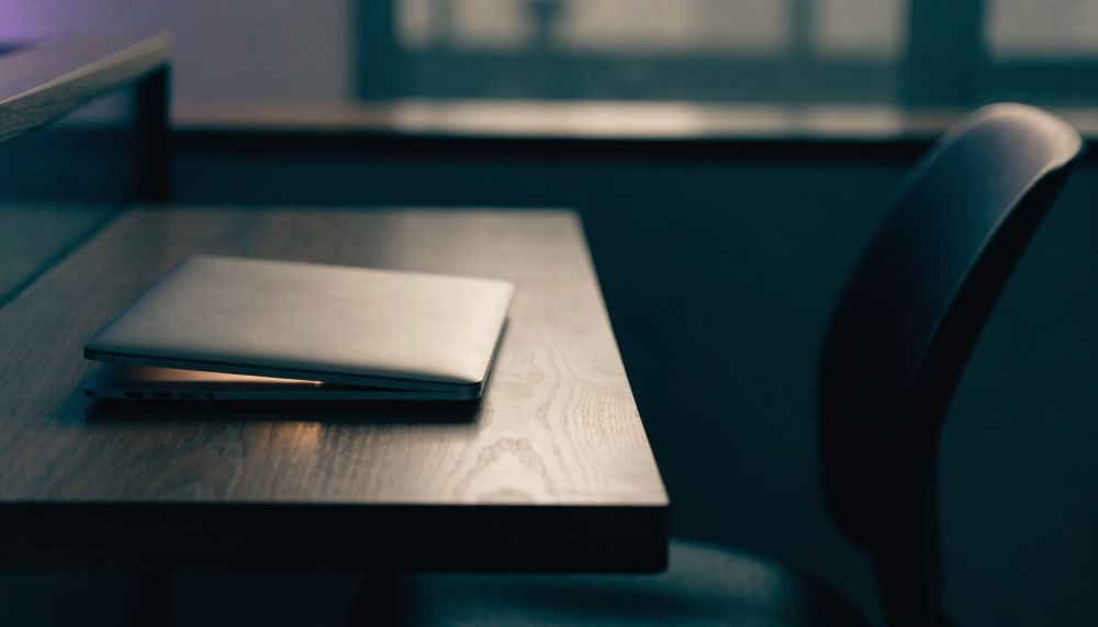A closed silver laptop resting on a wooden desk in a dim office after working hours, a thin sliver of warm light escapin