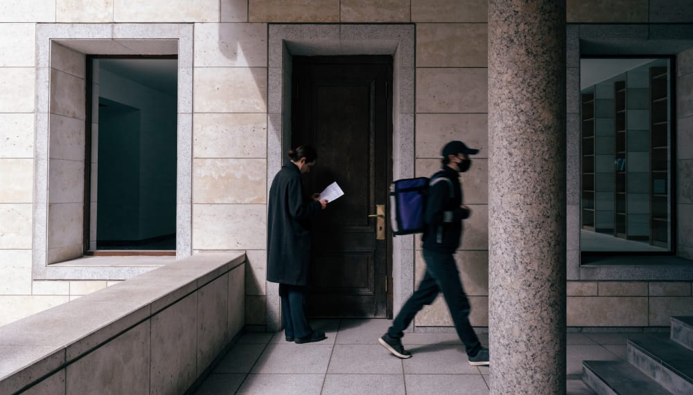 A closed doorway with a second figure standing beside it, reading a handwritten note before letting a courier pass. Quie