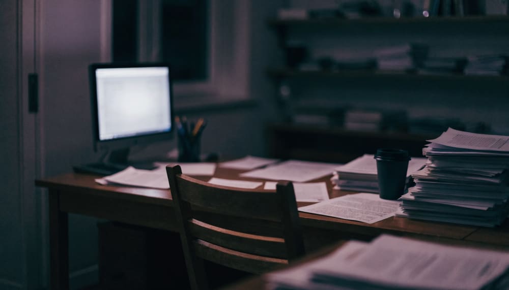 A dimly lit university office late at night, one monitor glowing on a cluttered wooden desk beside stacks of academic pa