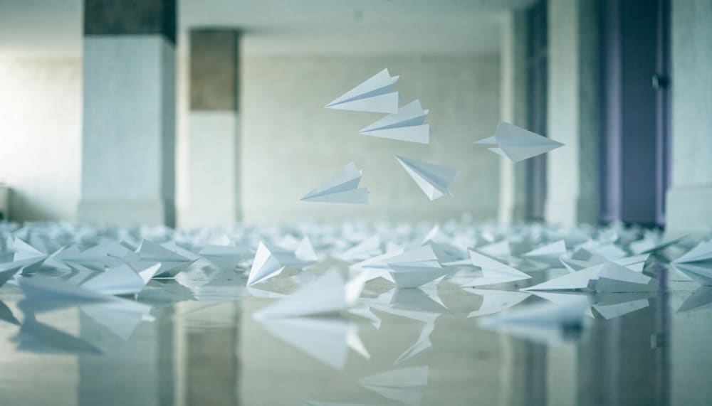 Hundreds of identical paper airplanes thrown across a bright empty exhibition hall, most scattered and falling short acr