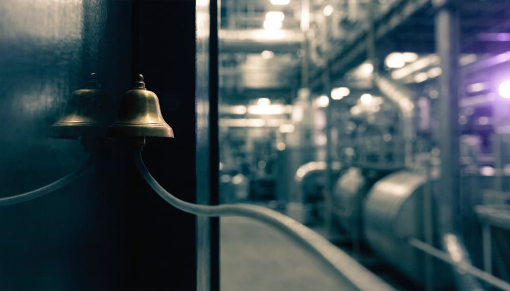 A tiny brass tripwire bell mounted beside a doorway, connected by a single thin cable running across the frame into a la