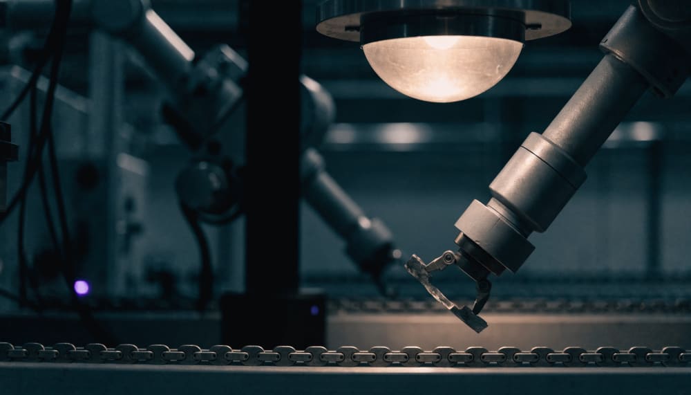 A long night-shift factory conveyor where a single robotic arm picks up an object, holds it under a lamp to inspect it, 