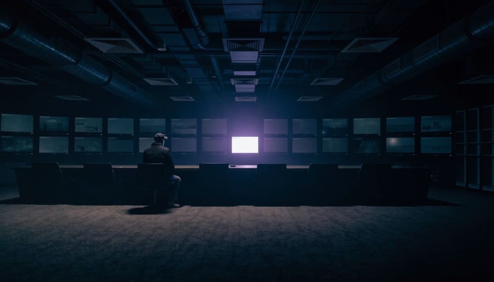 A wide control room at night where one operator faces a bank of identical dim monitors, with a single screen in the midd