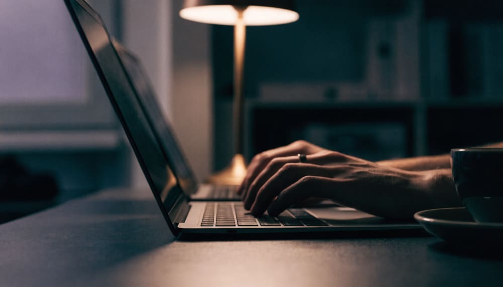 A developer workspace at night with two laptops open side by side running different coding assistants, hands mid keystro