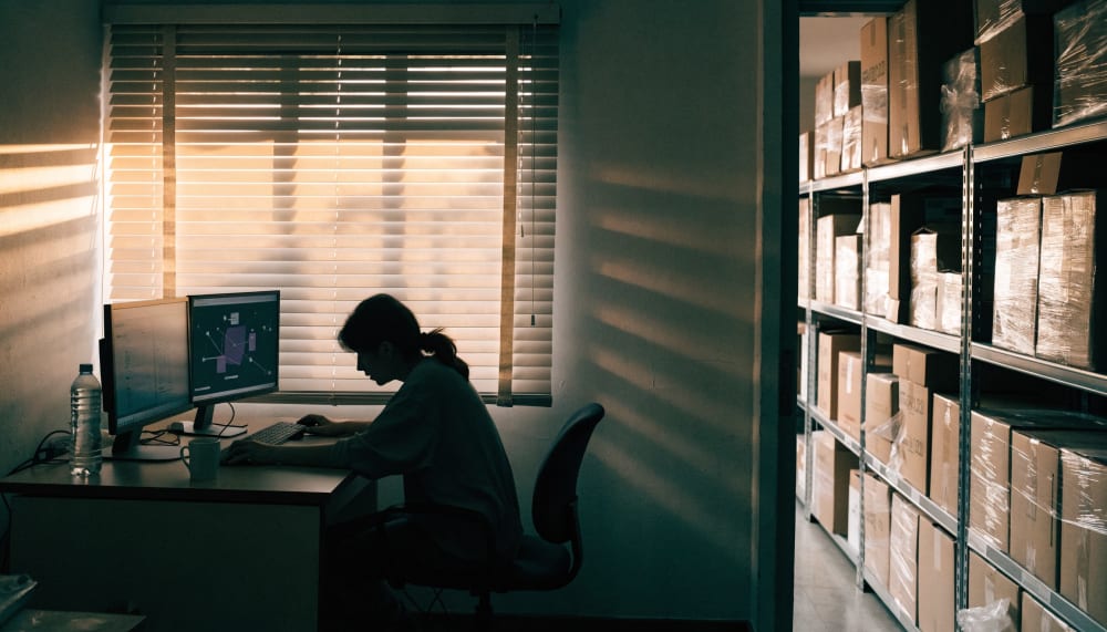 A small import company office in an Israeli industrial zone, storage shelves visible through an open doorway, one employ