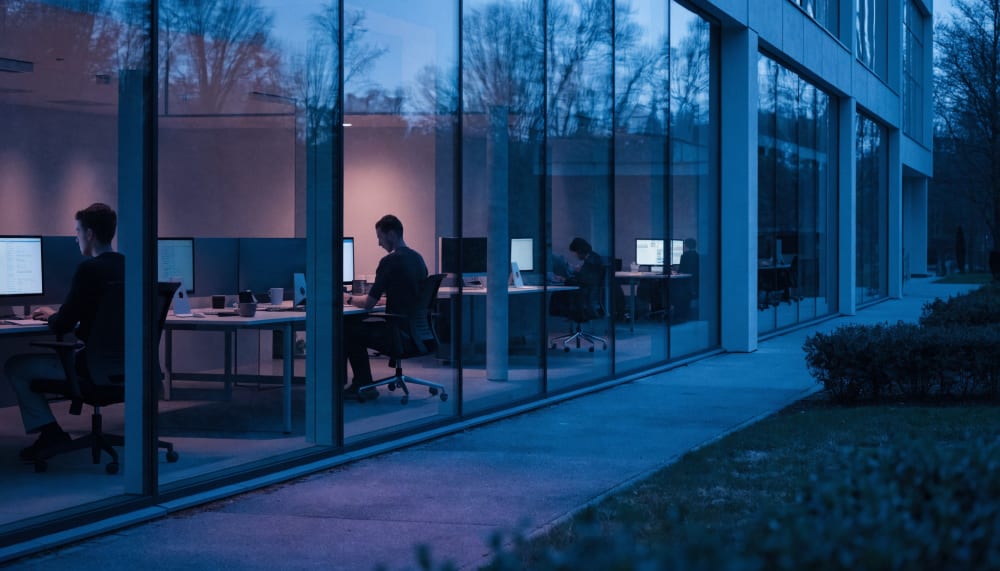 A quiet Stockholm office at dusk seen from outside through tall windows, a handful of people still at their desks, scree