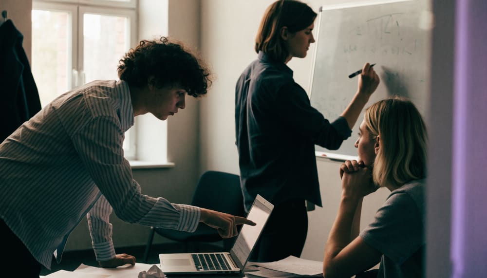 A small Israeli office team gathered around one laptop, someone pointing at the screen while another writes on a whitebo