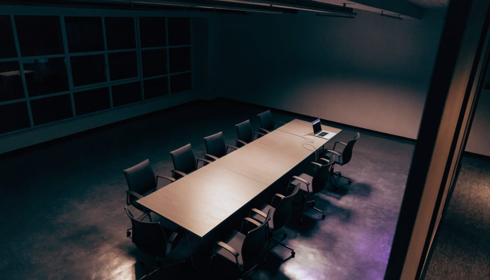 An empty conference room photographed from a high corner at night, chairs pushed back from a long table, one laptop stil