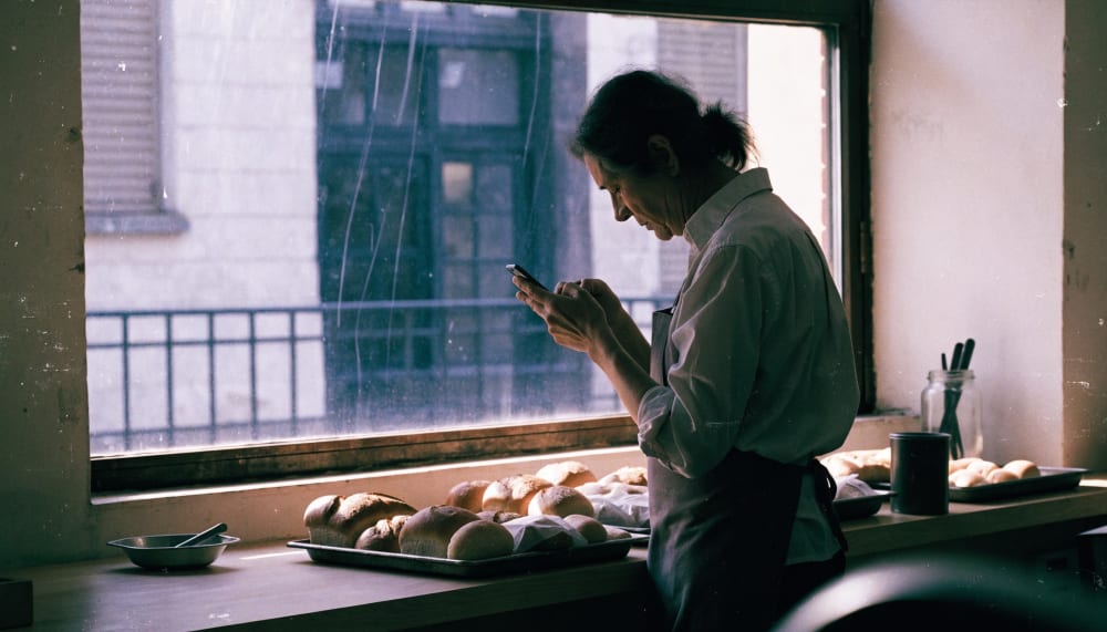 A small bakery owner standing behind the counter in the early morning, checking a phone, warm natural light through the 