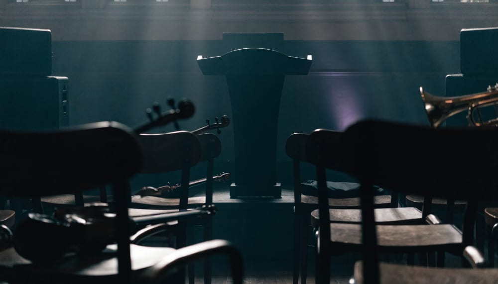 An empty orchestra conductor's podium seen from the musicians' seats, instruments resting on chairs in a dim hall, a sen