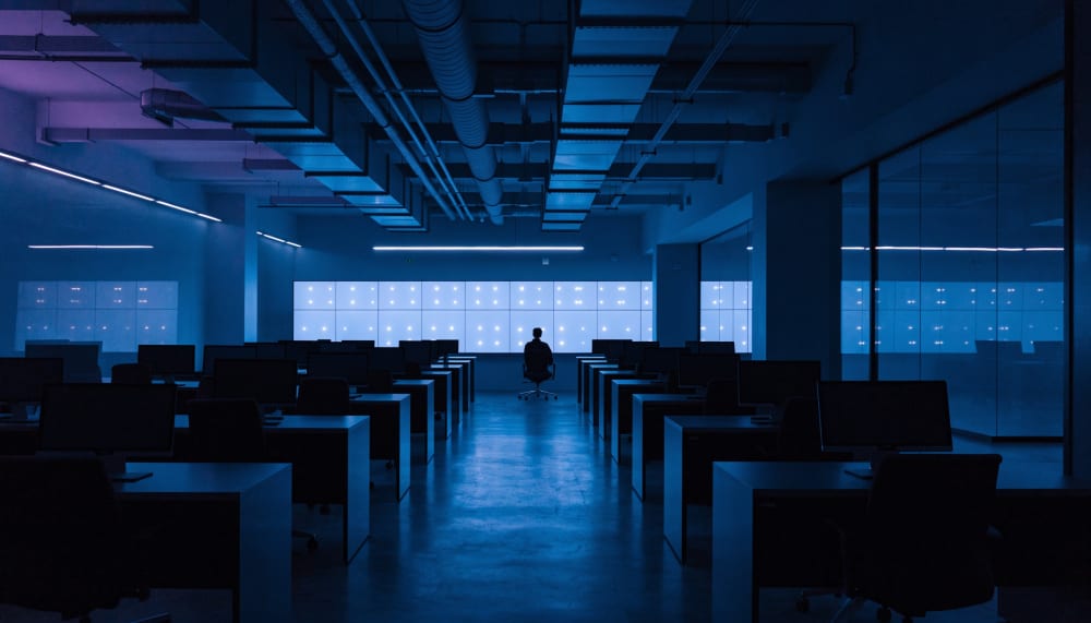 A dimly lit security operations centre at night, a single analyst seated at a desk facing a wall of glowing monitors, al