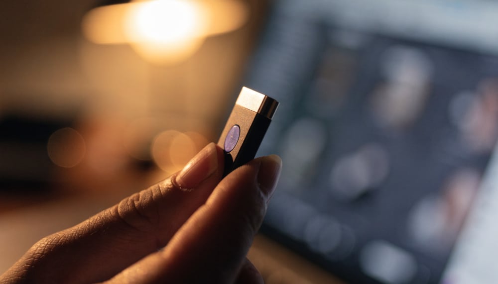 A close-up of a hardware security key held between two fingers in front of a blurred laptop screen, shallow depth of fie